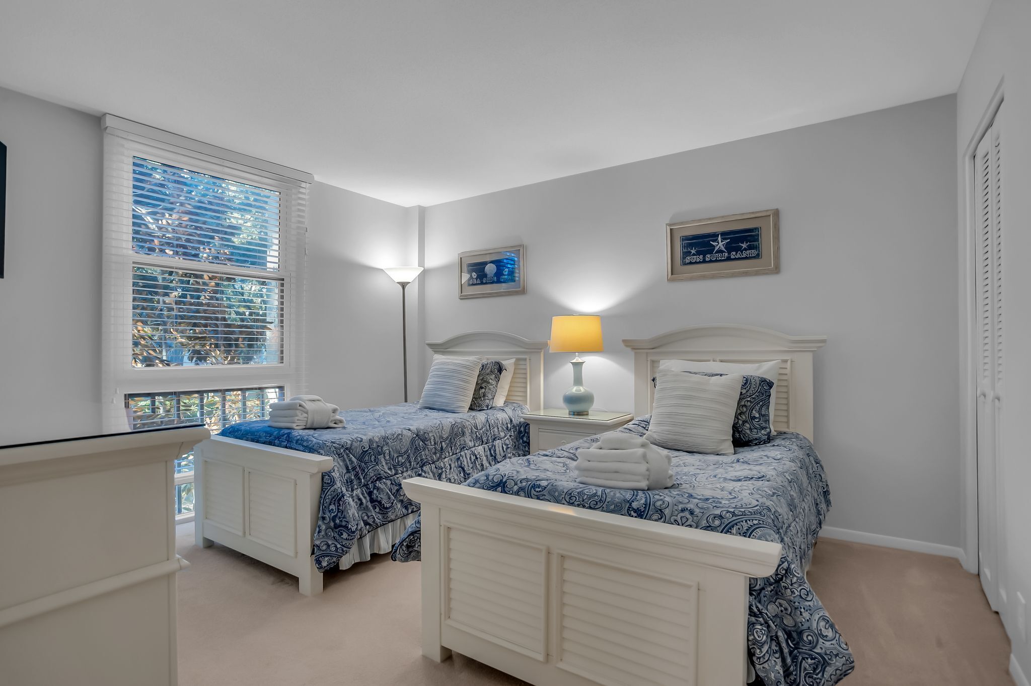 A bedroom with two beds, a window with blinds, a lamp, and framed pictures on the wall.