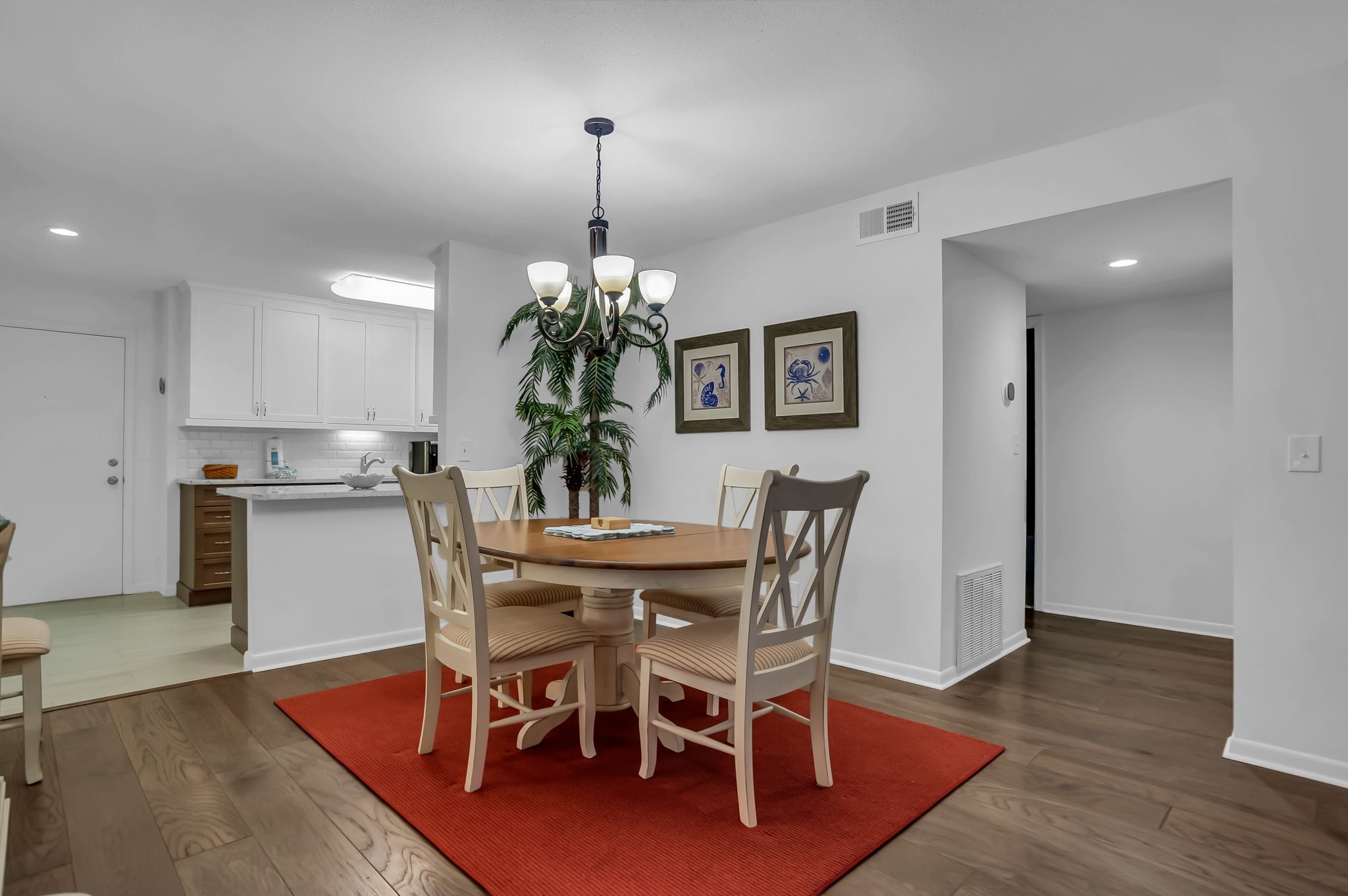 A dining area with a round table, four chairs, a red rug, a chandelier, and a kitchen in the background.