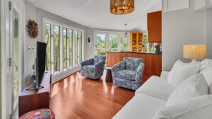 A spacious living room with a white sofa, patterned armchairs, a TV on a wooden stand, and a kitchen area with wooden cabinets and a hanging light fixture.