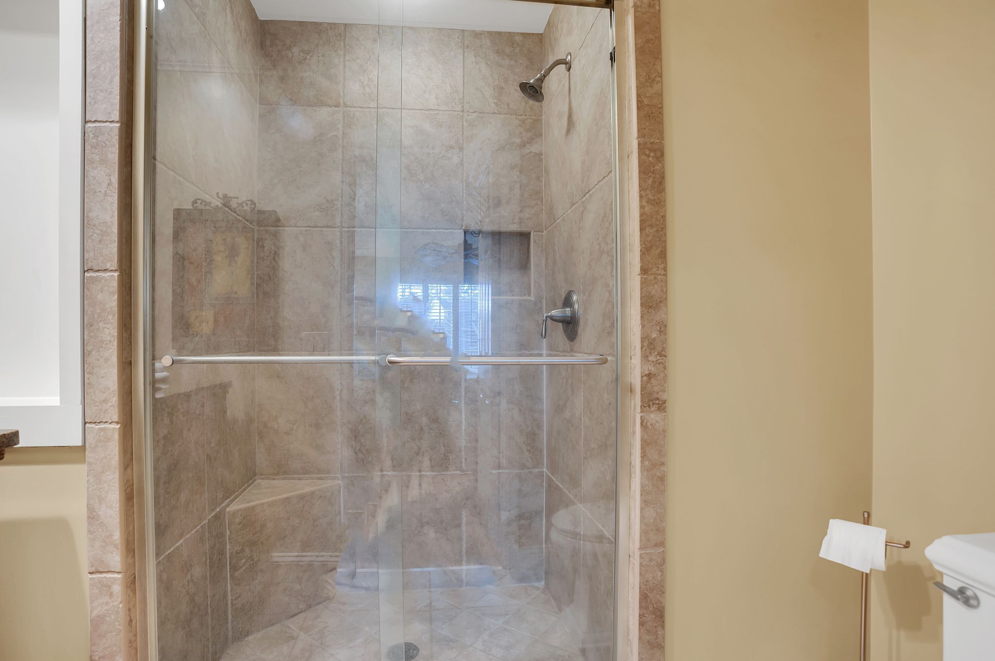 A glass-enclosed shower stall with a handrail, beige tiled walls, and a visible window.