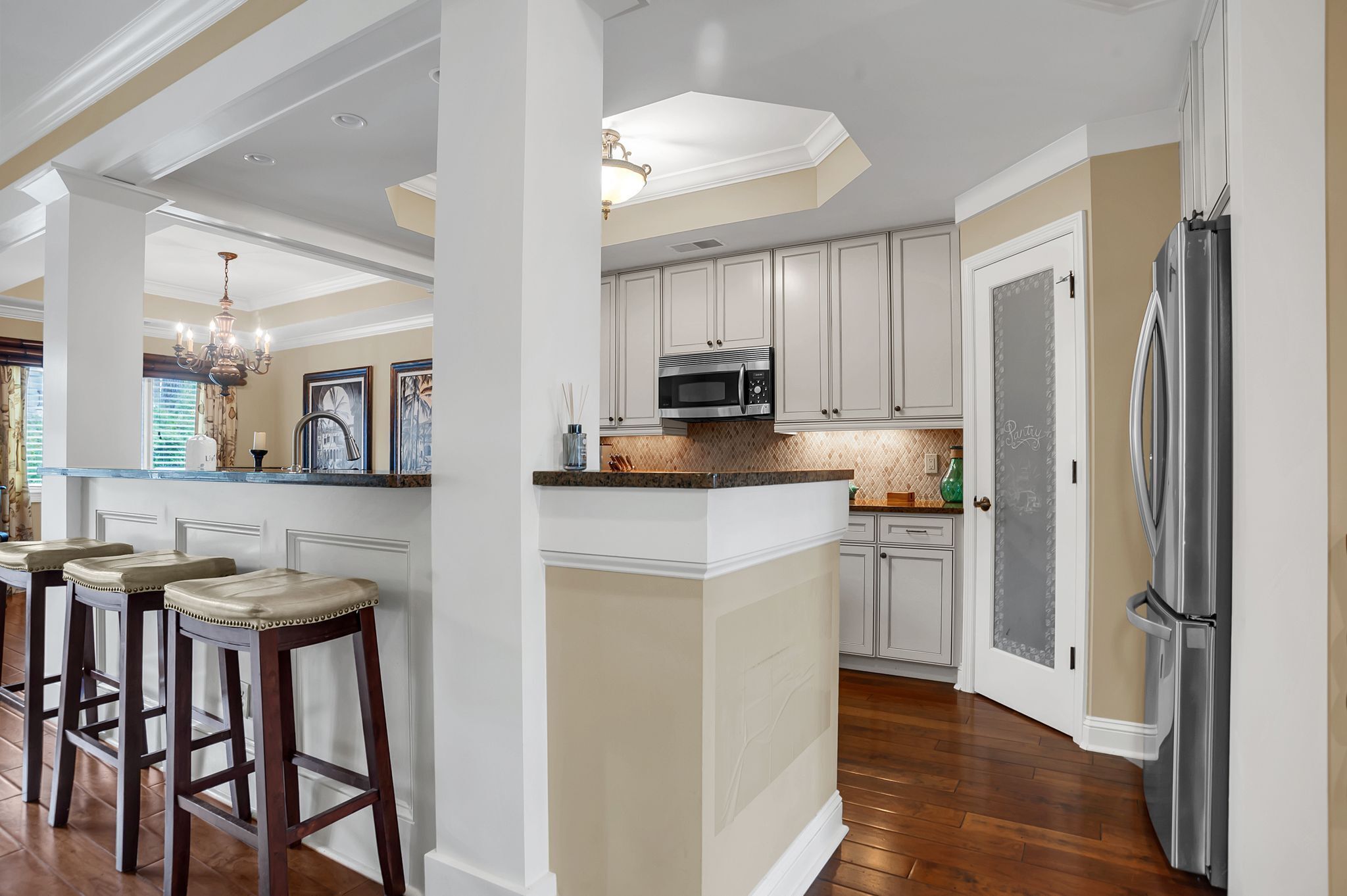 A kitchen with white cabinets, a central island with stools, a stainless steel refrigerator, and a chandelier.