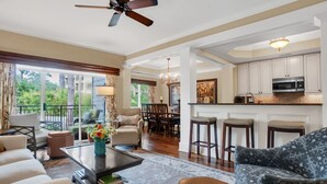 A spacious living room with a large window, a ceiling fan, a dining area with a chandelier, and a kitchen with bar stools. - 1112 Harbour South Club Villa at The Sea Pines Resort (Hilton Head Island)