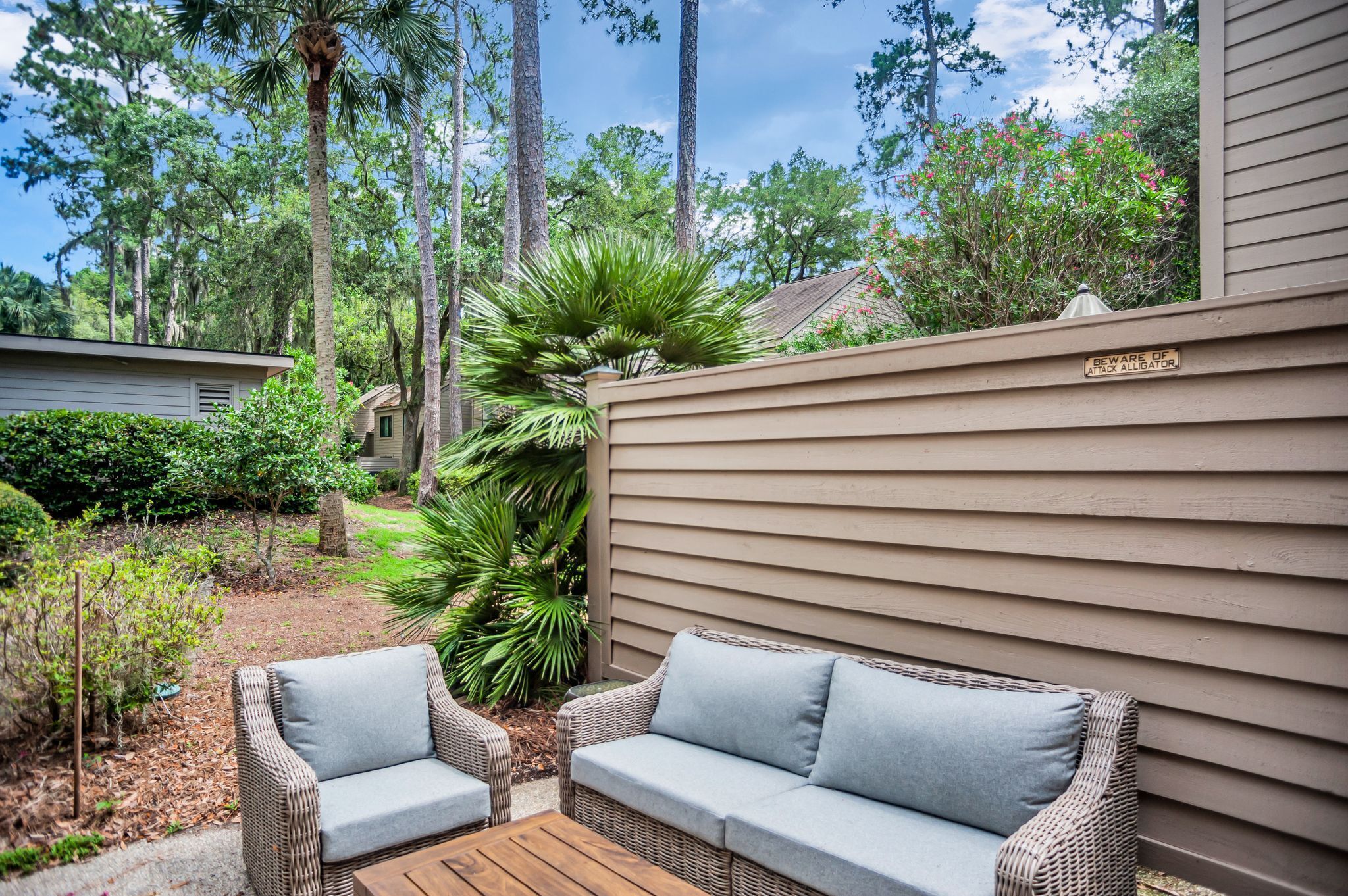 A patio area with wicker furniture and a wooden table, surrounded by lush greenery and tall trees.