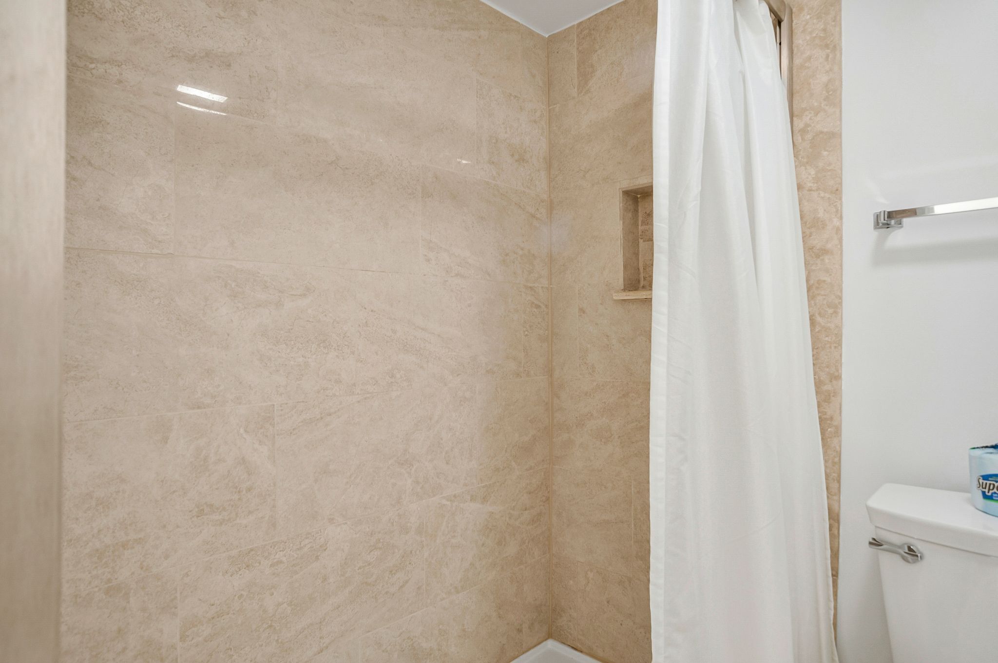 A bathroom with beige marble walls, a white shower curtain, and a toilet with a blue and white toilet paper holder.