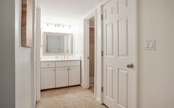 Un pasillo estrecho con una puerta blanca a la derecha, otra blanca a la izquierda y un baño con lavamanos y espejo blancos.