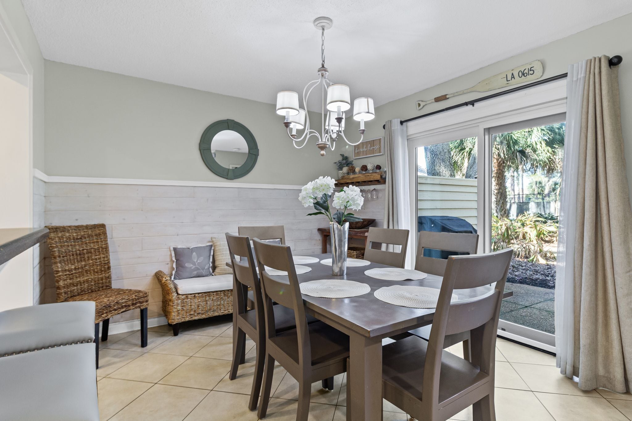 Un coin repas avec une table dressée pour quatre personnes, entouré de chaises en osier et d’un banc rembourré. Un lustre est suspendu au plafond, et un miroir est fixé au mur. Une porte-fenêtre coulissante donne sur un espace extérieur agrémenté de plantes.