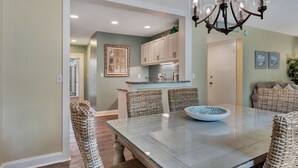 A dining area with a large table, wicker chairs, a chandelier, and a kitchen in the background.