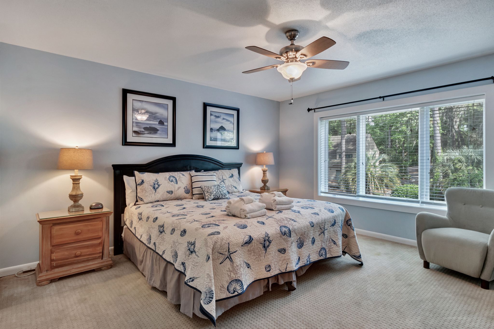 A bedroom with a bed, two bedside lamps, a ceiling fan, a window with blinds, and two framed pictures on the wall.