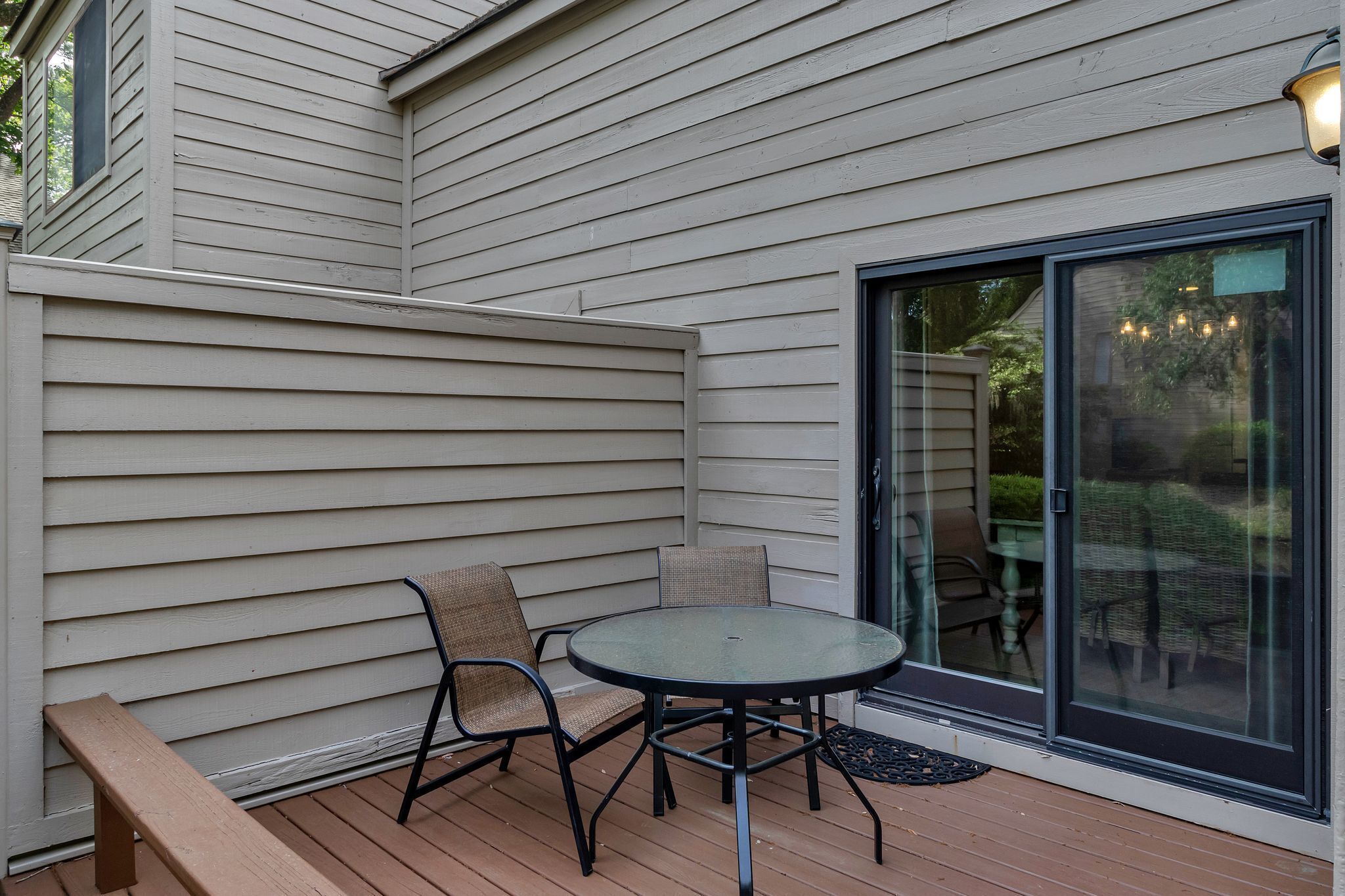 A covered patio with a round table and two chairs, a wooden bench, and a sliding glass door leading to a garden area.