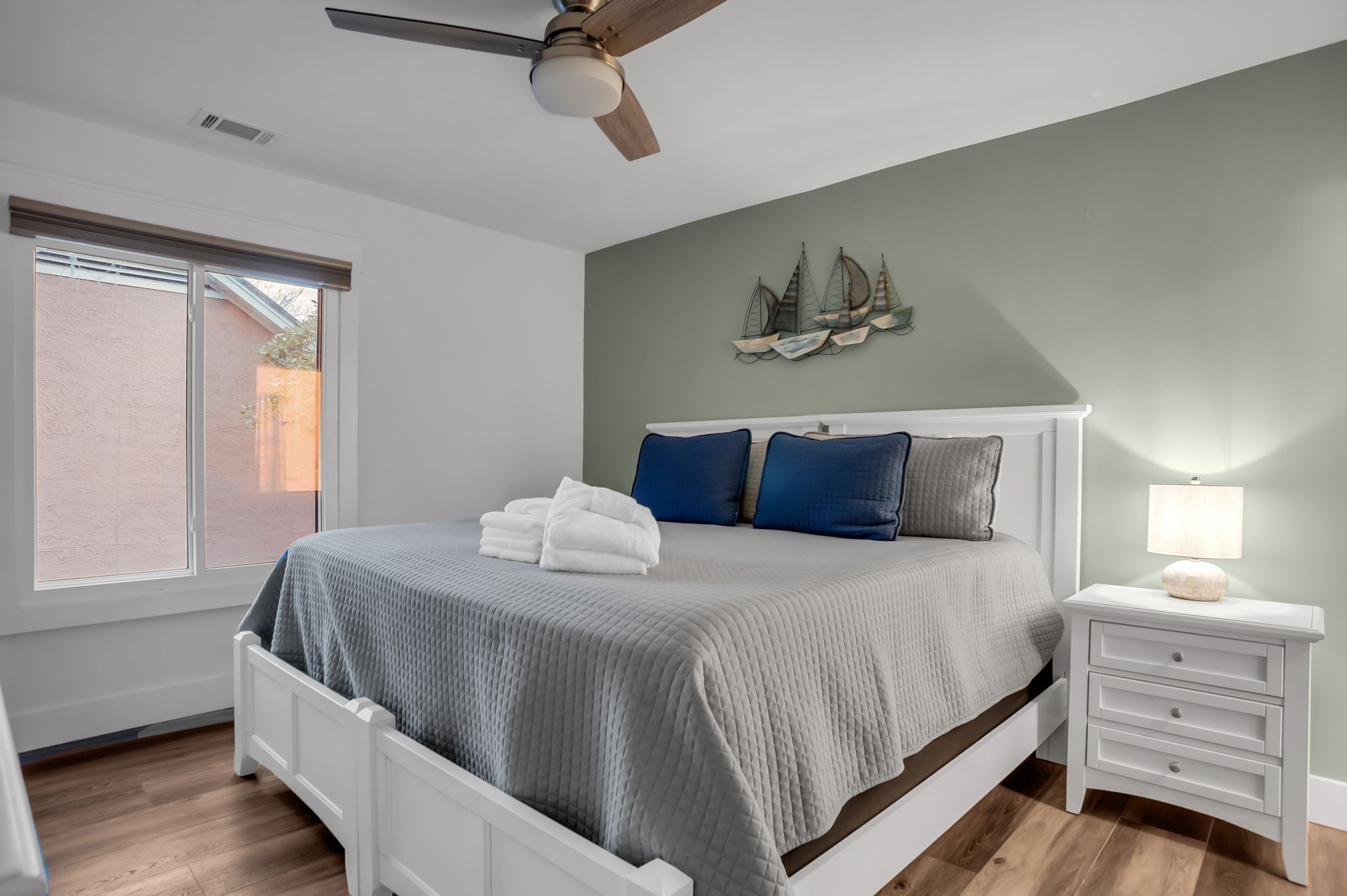 Une chambre à coucher comprenant un lit, une table de chevet, un ventilateur de plafond, une fenêtre et un élément décoratif mural.