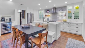 A modern kitchen with white cabinetry, a central island, and a dining area with wooden chairs. - 26 Muirfield Road at The Sea Pines Resort (Hilton Head Island)