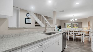 Une cuisine moderne avec des meubles blancs, un comptoir en granit et un évier en acier inoxydable. On y trouve un coin repas avec une table et des chaises en bois, ainsi qu’un escalier menant à un autre niveau.