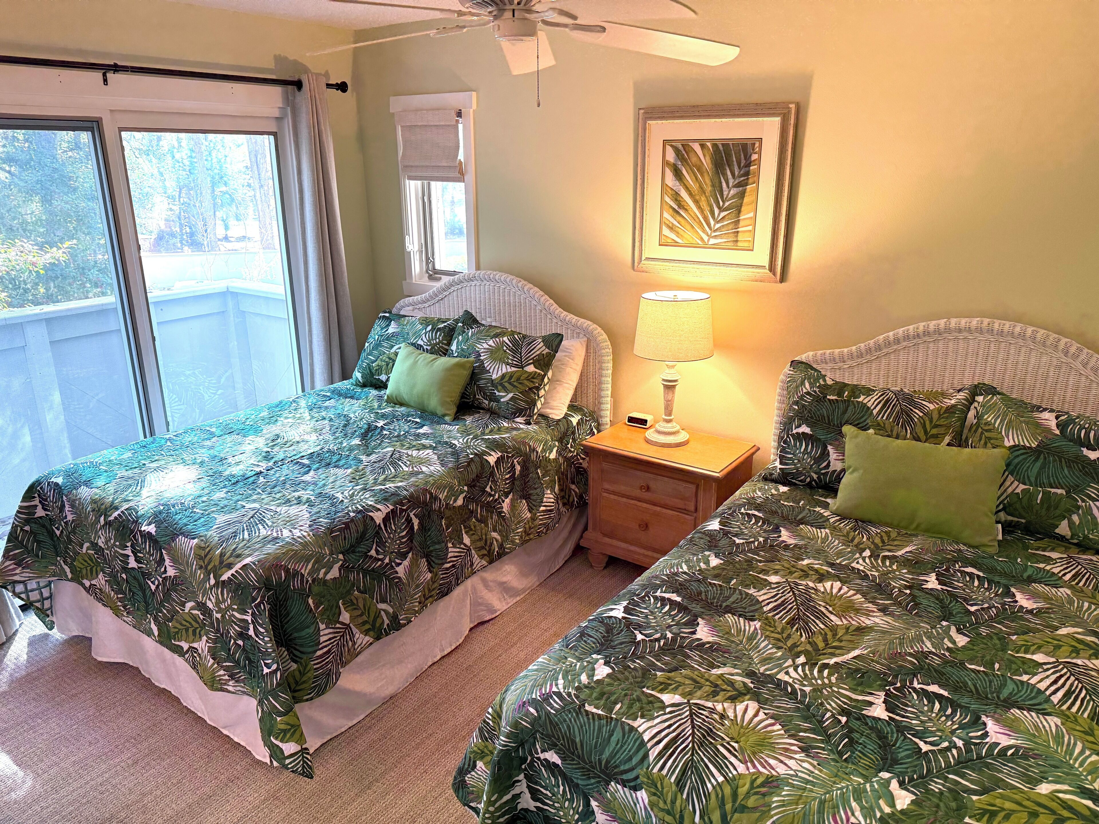 A bedroom with two beds, a nightstand with a lamp, a ceiling fan, and a window with blinds.