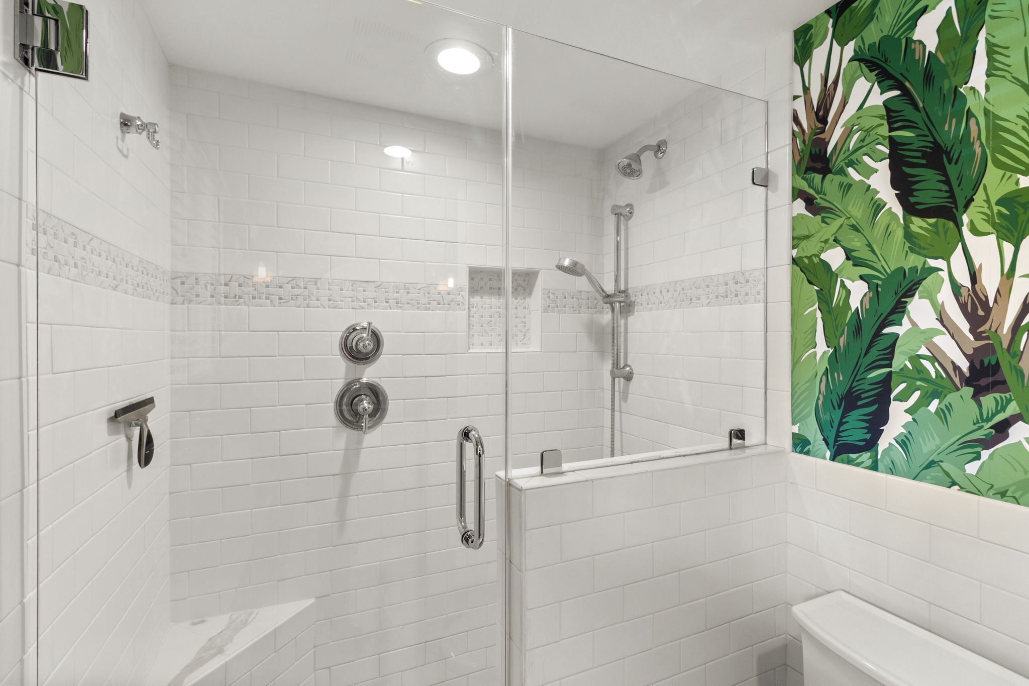 A modern bathroom with white tiles, a glass shower enclosure, and a green leaf-patterned wall.
