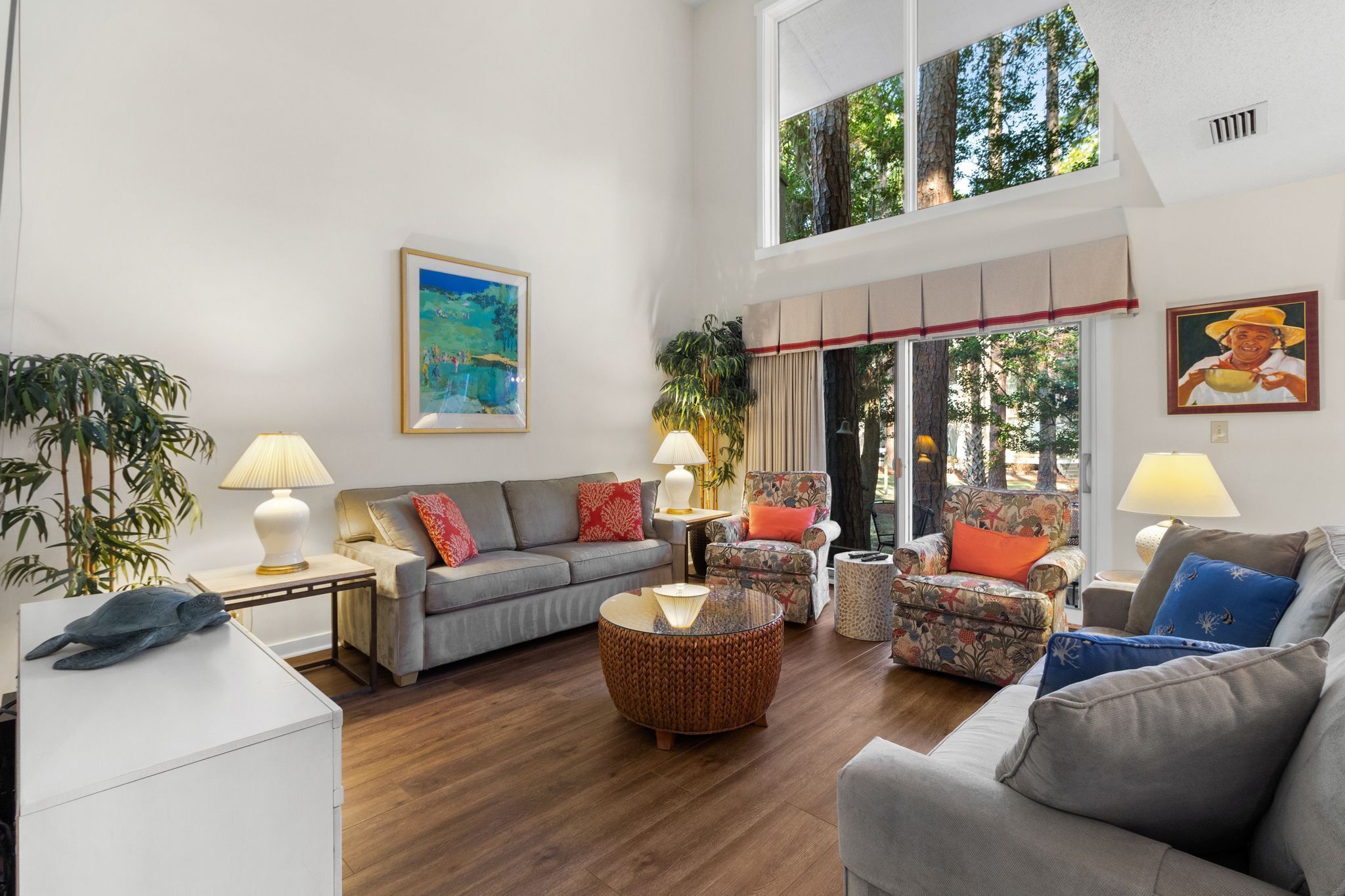 A spacious living room with a grey sofa, a round coffee table, and a floral armchair. There are two lamps on side tables, and a painting on the wall.