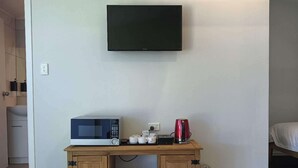 Una pequeña cocina equipada con microondas, tostadora y cafetera sobre un mueble de madera, un televisor montado en la pared y una cama al fondo.