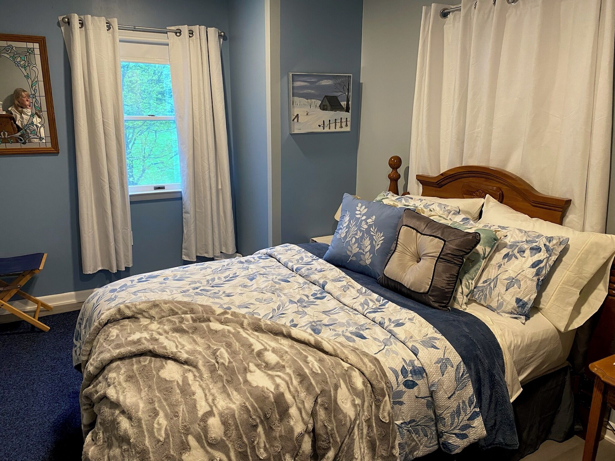 Un dormitorio con cama, cortinas, una ventana, un cuadro en la pared y una mesita.