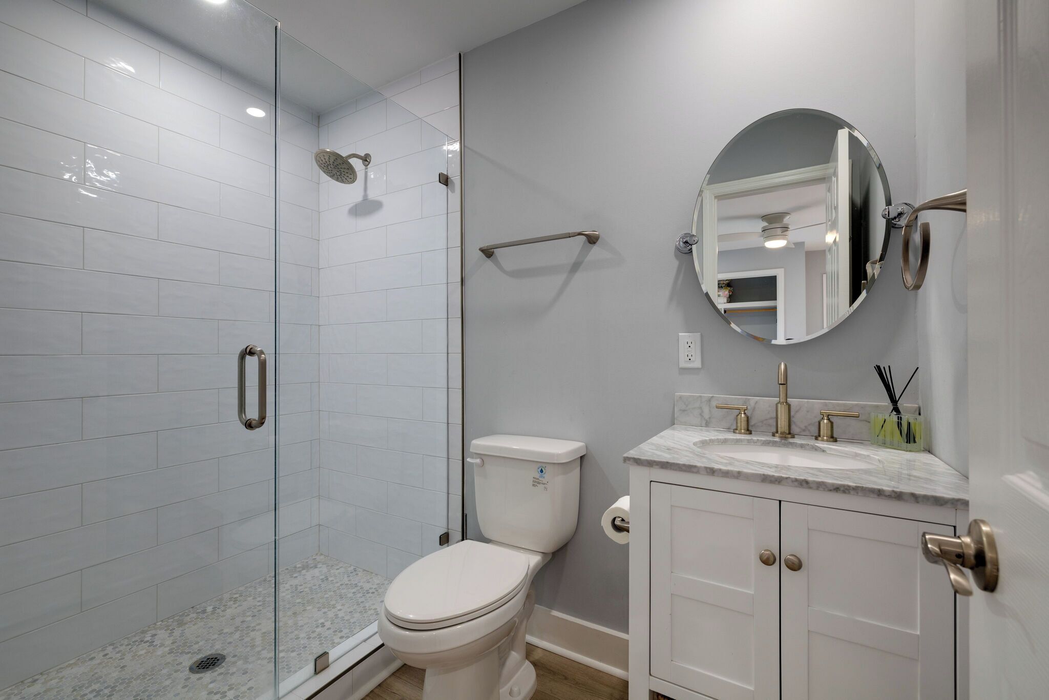 A modern bathroom with a glass-enclosed shower, a white toilet, a marble sink, and a round mirror.