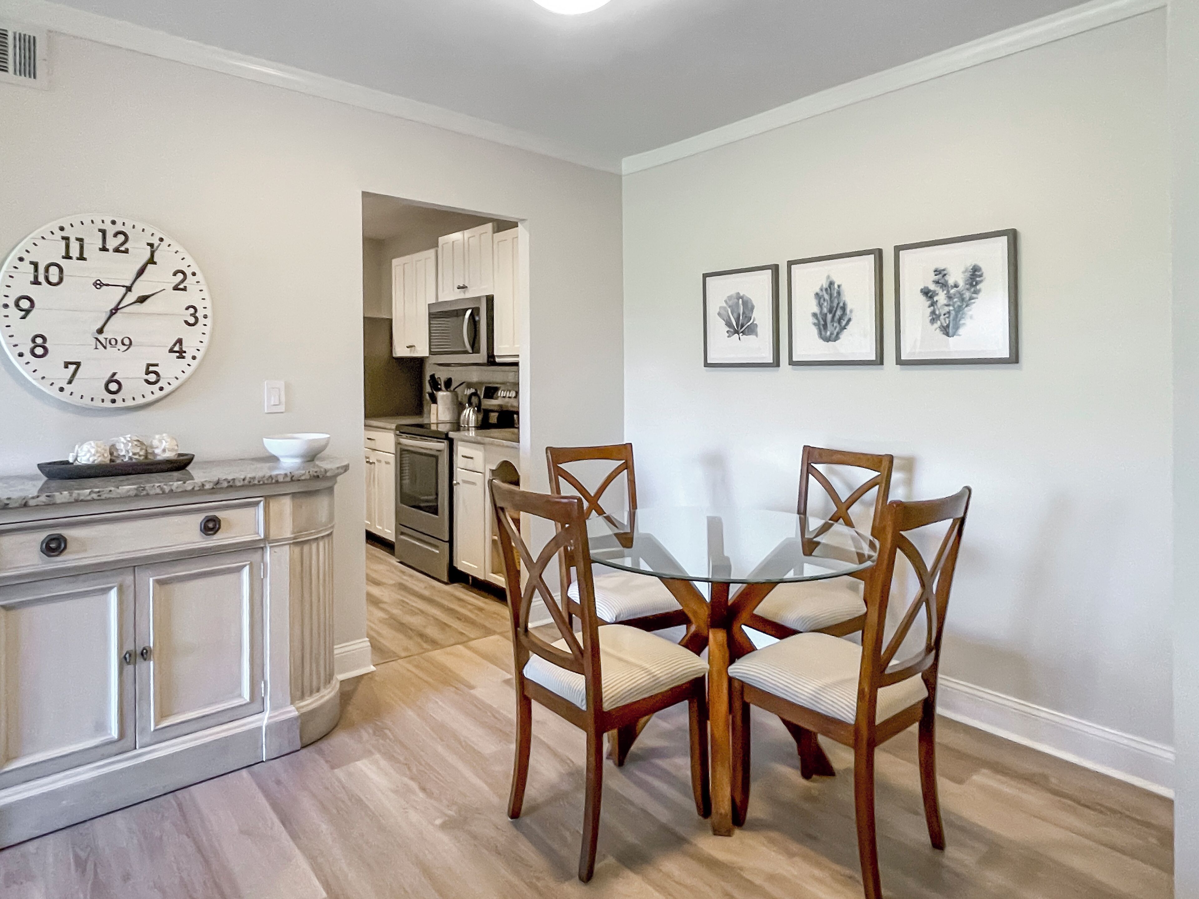A dining area with a round glass table and wooden chairs, a kitchen in the background, and a wall clock.