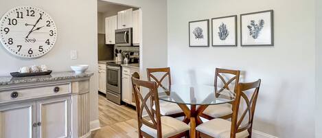A dining area with a round glass table and wooden chairs, a kitchen in the background, and a wall clock.