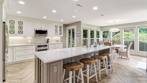 A spacious kitchen with white cabinetry, a central island with stools, and large windows offering a view of greenery.