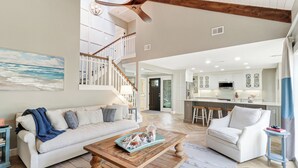 A spacious living room with a high ceiling, a wooden coffee table, a white sofa, and a view of the kitchen and dining area.