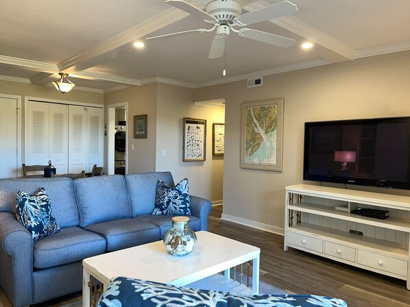 A living room with a grey sofa, a white coffee table, a TV on a white stand, and a ceiling fan. - 1797 Bluff Villas at The Sea Pines Resort (Hilton Head Island)