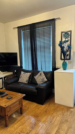 A living room with a black leather sofa, two patterned throw pillows, a wooden coffee table, a white side table, a flat-screen TV, and a framed floral artwork on the wall. - 2 Bedroom House in Manchester (Failsworth)