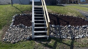 A house with a wooden staircase leading to the front door, surrounded by a garden with a stone border. - This Quaint home overlooking Cayuga Lake is the perfect Finger Lakes Getaway! (Seneca Falls)