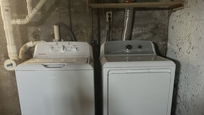 Two white front-loading washing machines in a laundry area with visible pipes and a wooden shelf above. - This Quaint home overlooking Cayuga Lake is the perfect Finger Lakes Getaway! (Seneca Falls)