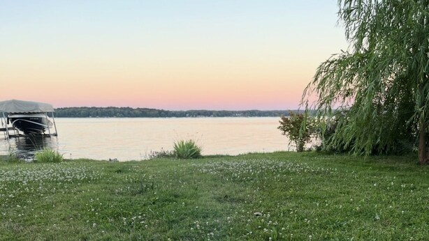 A circular fire pit with logs burning, surrounded by a stone border, set against a lakeside backdrop. - This Quaint home overlooking Cayuga Lake is the perfect Finger Lakes Getaway! (Seneca Falls)