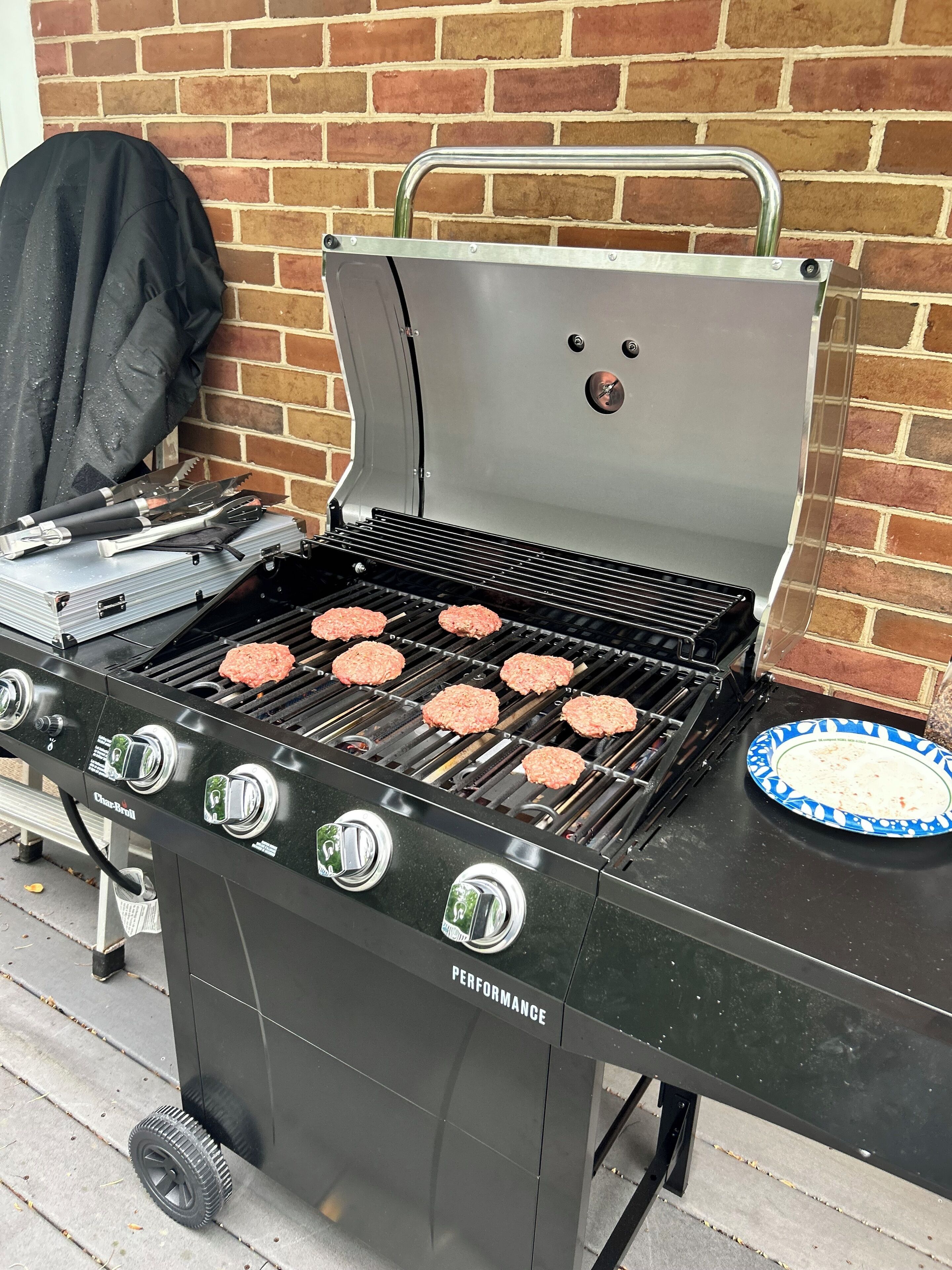Ein Char-Broil Grill mit vier Bratburgern, einer Grillbürste und einem Papierteller daneben.
