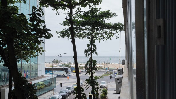 Une vue depuis une fenêtre donnant sur une rue animée, avec des voitures, une plage et un bâtiment moderne aux façades vitrées.
