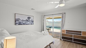 A bedroom with a bed, a ceiling fan, a bedside lamp, a large window with curtains, a view of a pool, and a wooden bench.