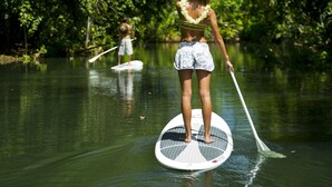 Duas pessoas praticam stand-up paddleboarding em um rio calmo, cercadas por vegetação exuberante.