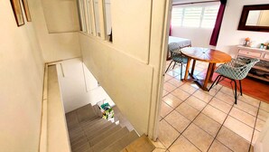 Uma escadaria leva a uma sala de jantar com mesa e cadeiras, uma área de cozinha com armários e uma janela com persianas.