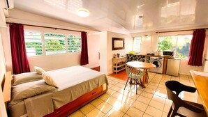 Quarto de hotel compacto com cama, pequena mesa de jantar, área de cozinha com máquina de lavar e vista para vegetação pelas janelas.