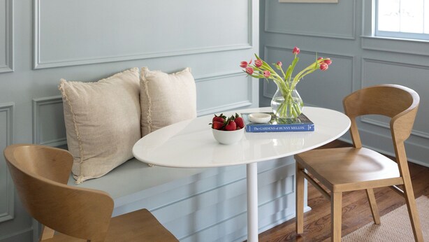 Una habitación con una mesa redonda blanca, dos sillas de madera, un jarrón con flores y un cuadro en la pared.