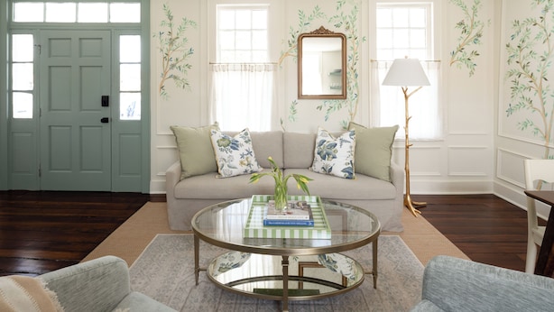 Una sala de estar con puerta verde, sofá con cojines estampados, mesa de centro de cristal y una lámpara de pie.