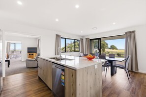 A modern kitchen with a central island, a dining area with a table and chairs, a built-in dishwasher, and large windows offering a view of the outdoors. - Tranquil and Unique In A Gated Grandview Estate Community With Amazing Views (Taupō)