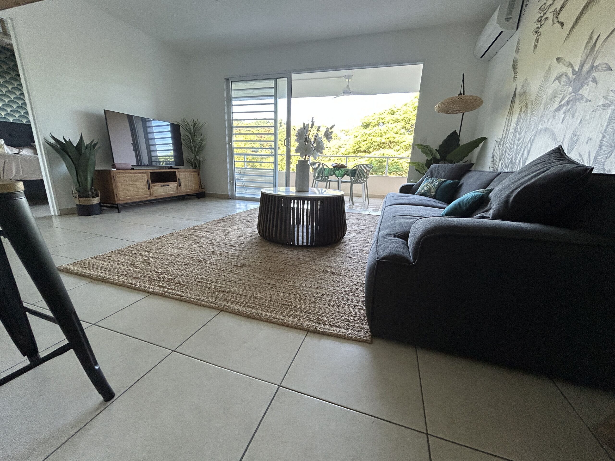 Una sala de estar moderna con un sofá gris, una mesa de centro redonda y un televisor sobre un mueble de madera. Se ve una ventana grande con vistas a zonas verdes.