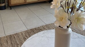 Ένα σαλόνι με μια τηλεόραση τοποθετημένη πάνω σε ένα ξύλινο έπιπλο, ένα μεγάλο παράθυρο και ένα βάζο με λευκά λουλούδια.
