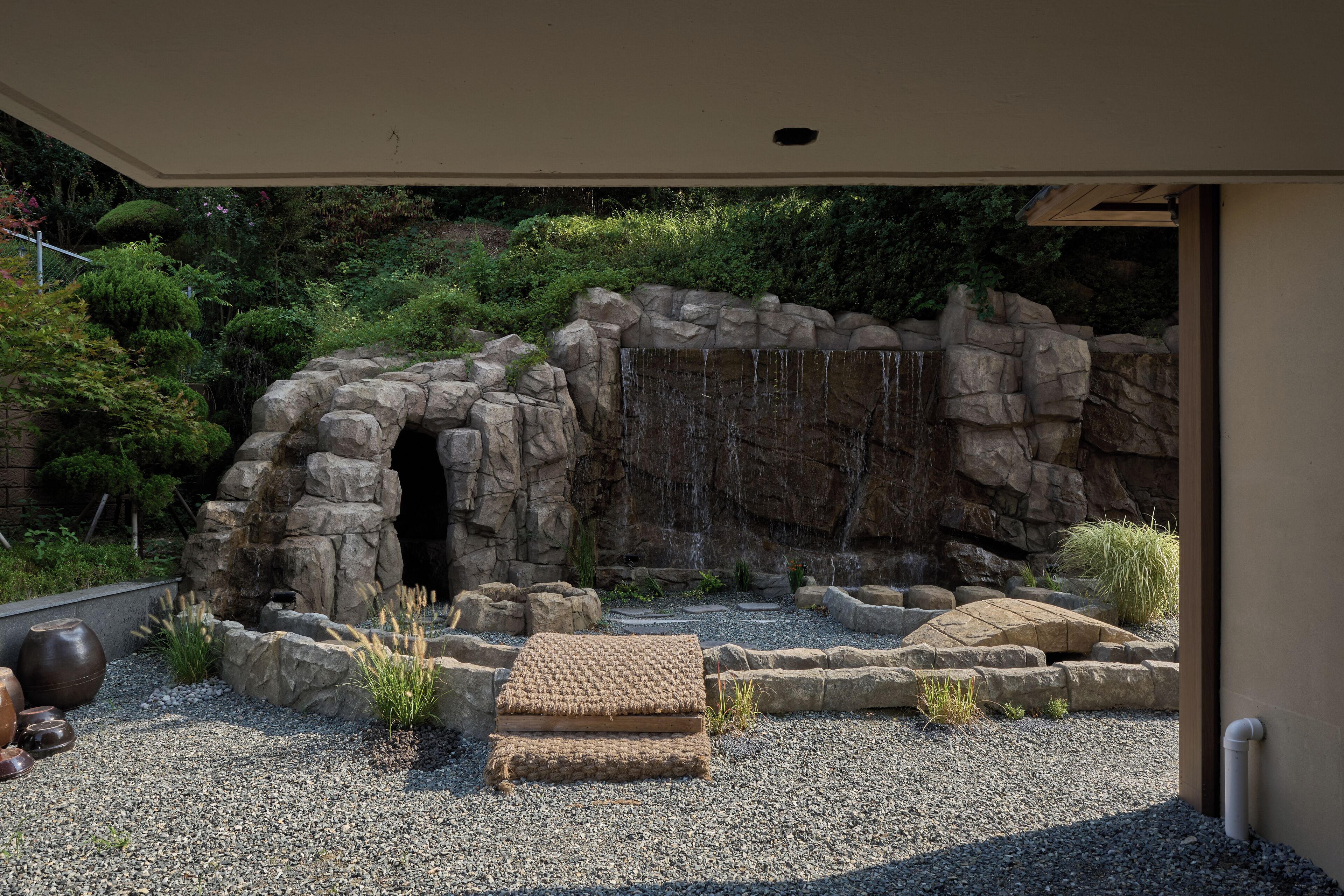 A rock garden with a waterfall, surrounded by a stone archway and various plants.