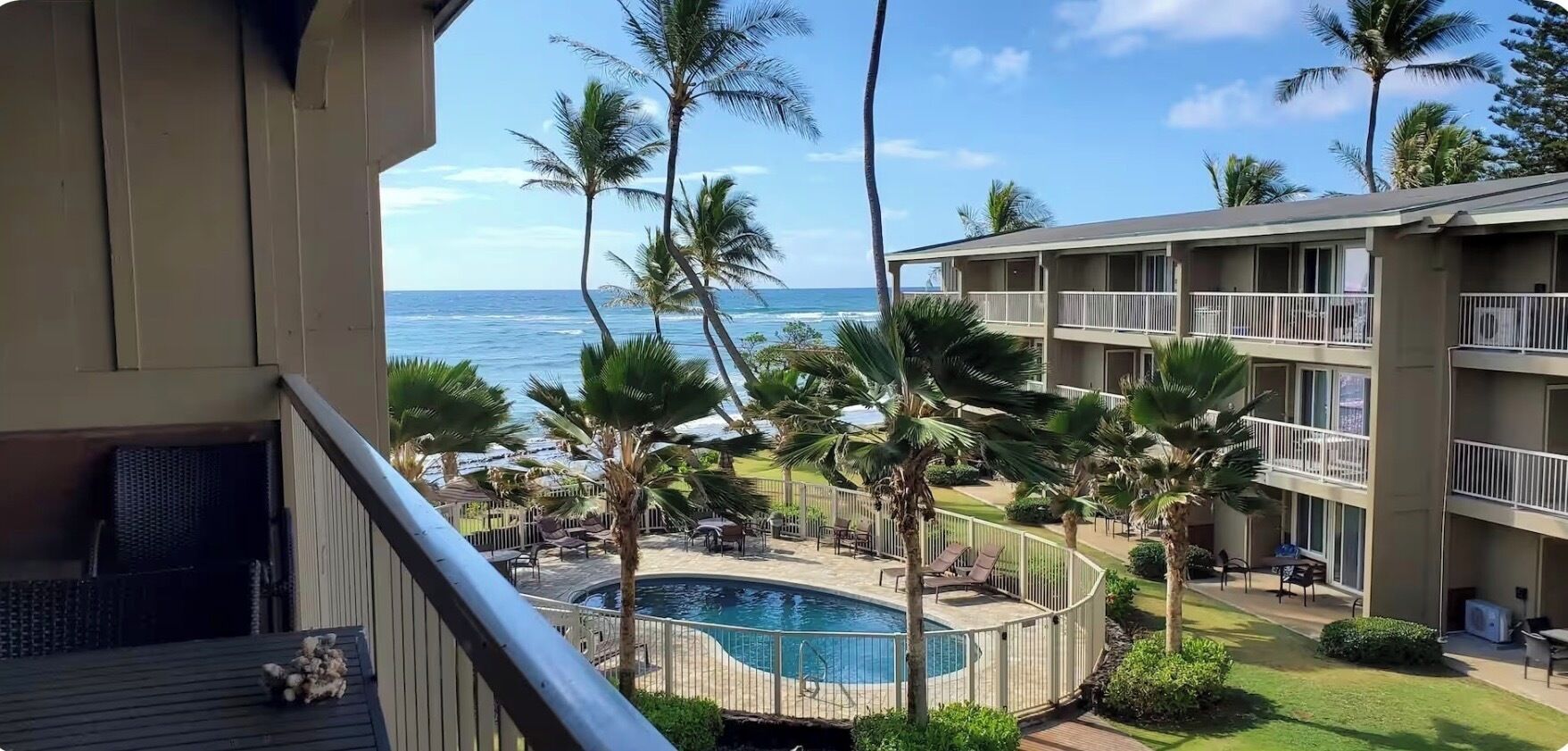 A pool area with lounge chairs and palm trees, overlooking a body of water and a building with balconies.