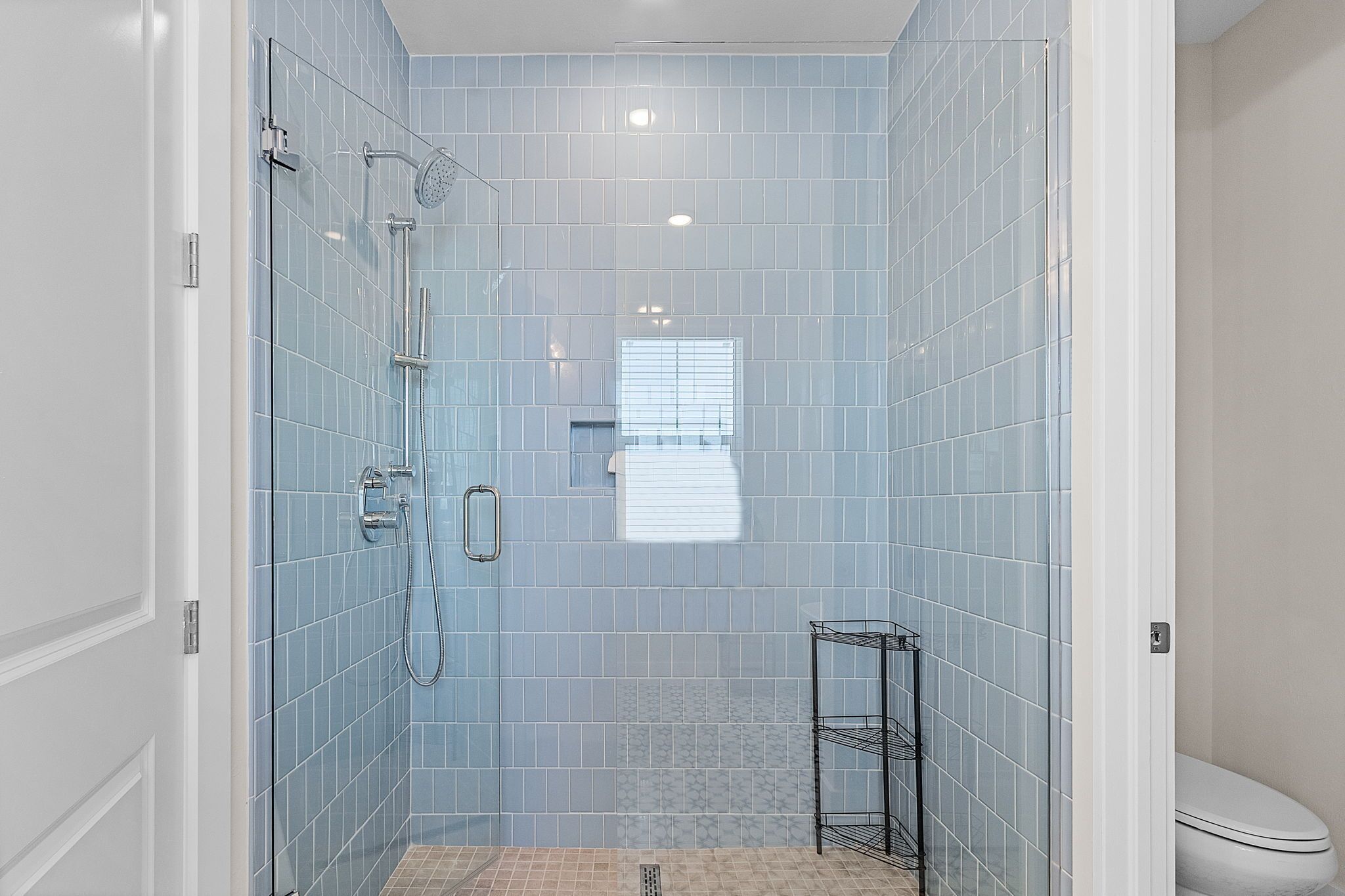 A bathroom with blue tiled walls, a glass shower enclosure, a toilet, and a small metal shelf.