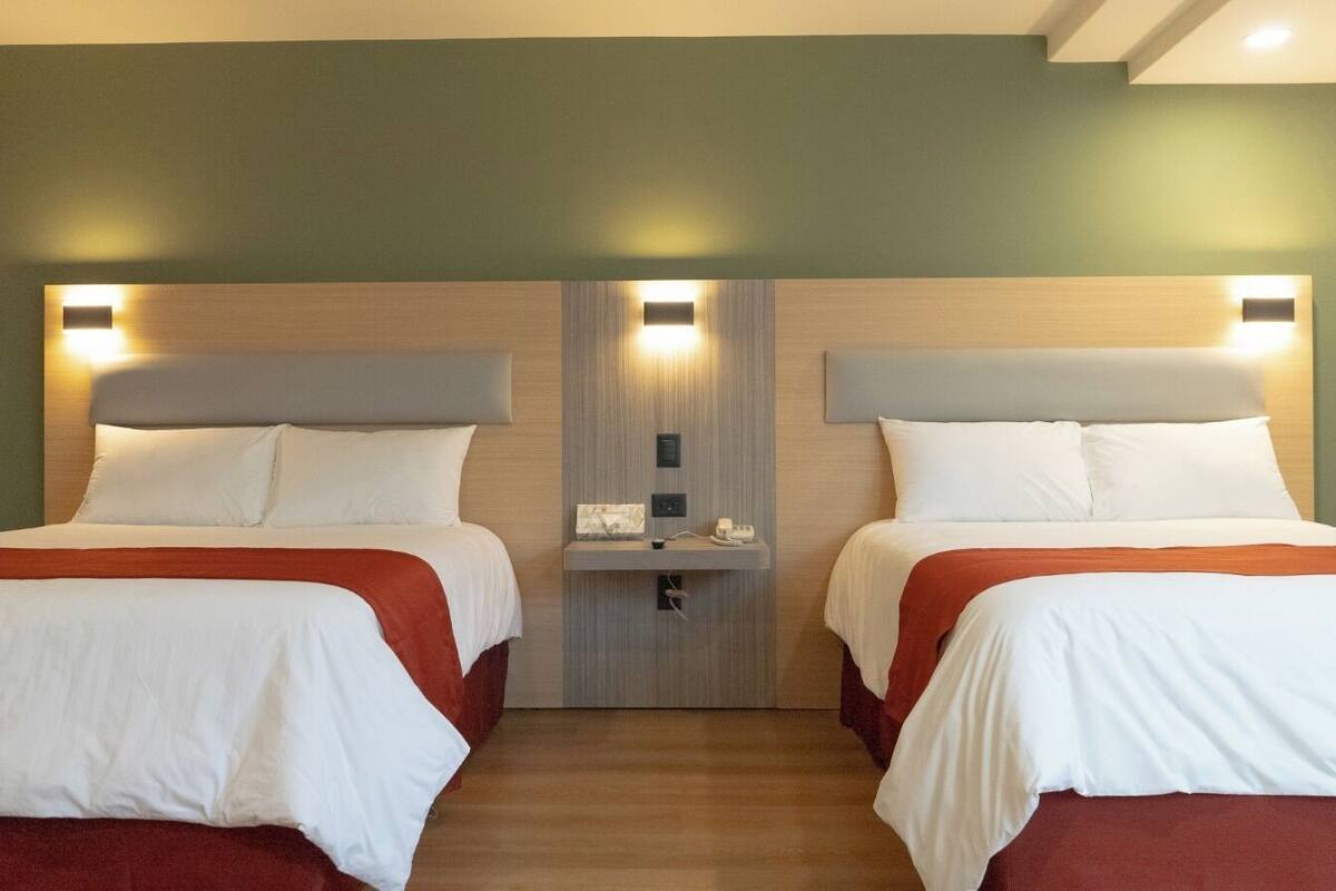 Une chambre d’hôtel avec deux lits, chacun recouvert de draps blancs et d’une couverture rouge. Il y a une petite table de chevet avec une lampe et un téléphone. La chambre a un plancher en bois et des murs verts.