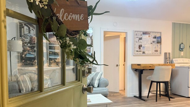 Una stanza con un cartello che dice 'Welcome', una porta in legno, un divano bianco, un tavolino, una sedia e un angolo cottura con frigorifero e un piano di lavoro in legno.