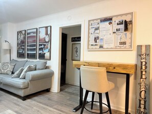 Una sala de estar con un sofá gris, un taburete de madera y un tablón de anuncios con varios avisos.