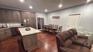 A spacious kitchen with a central island, a dining area with a table and chairs, and a brown leather sofa in a living area.