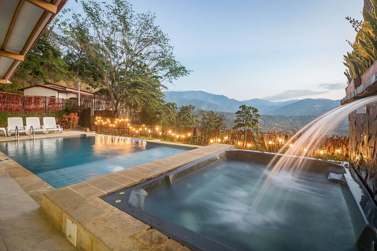Ein Pool mit Whirlpool und Wasserfall, umgeben von einem Bambuszaun und Lichterketten, mit einer Berglandschaft im Hintergrund.